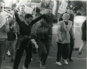 A black and photograph of an anti Apartheid rally. People walk together holding placards with their arms in the air. 