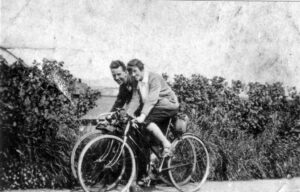 A black and white photograph of two cyclists in the early 1900s. 