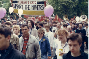 A group of people on a protest march during the Miners' strike 1984-1985. They hold pink balloons and a banner that states 'Lesbians and Gays Support the Miners'.